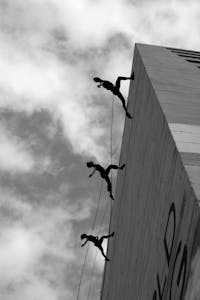 Three dancers perform a daring vertical dance on a building wall, silhouetted against the sky.